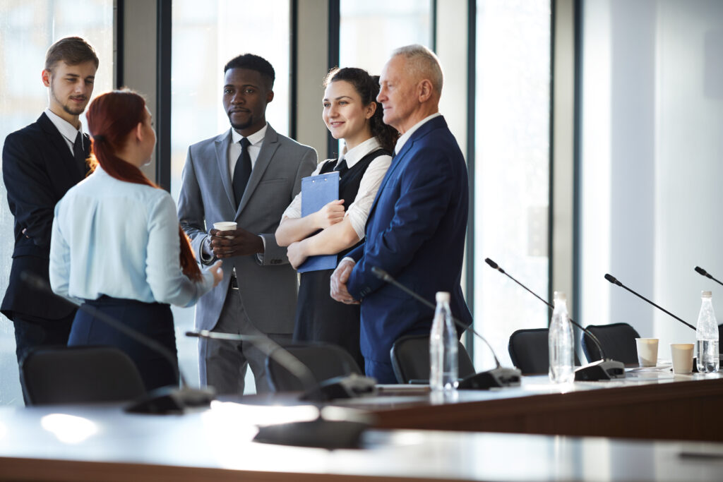 Five people in business attire stand and talk in a modern conference room with microphones, bottled water, and large windows in the background.